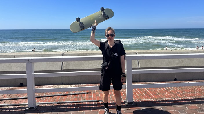 Mia Djuric steht auf der Promenade in Sylt und hält ihr Skateboard hoch.