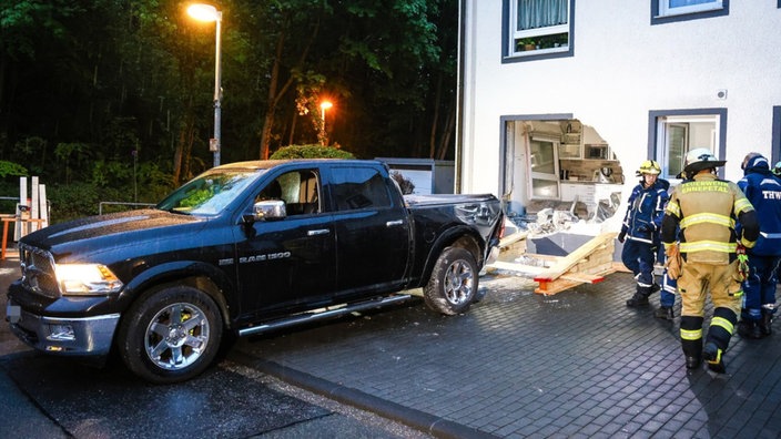 Ein Pick-up steht vor einem großen Loch in einer Hauswand, durch das Loch ist die Küche einer Wohnung zu sehen. Rechts stehen Einsatzkräfte von Feuerwehr und Technischem Hilfswerk