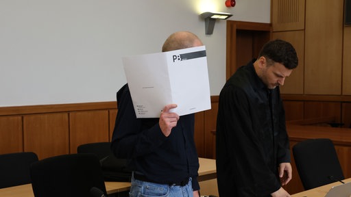 Ein vor Gericht angeklagter Mann hält sich in einem Gerichtssaal eine weiße Mappe vor das Gesicht. Neben ihm steht ein Mann in schwarzer Kleidung.