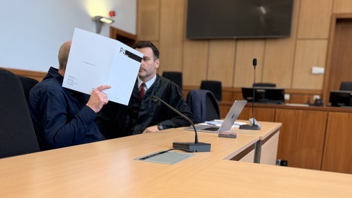 Ein Mann sitzt mit seinem Anwalt im Gerichtssal und hält sich eine Mappe vor das Gesicht.