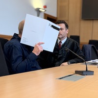 Ein Mann sitzt mit seinem Anwalt im Gerichtssal und hält sich eine Mappe vor das Gesicht.
