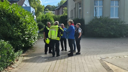 Eine Gruppe Männer, teilweise mit gelben Westen und der Aufschrift stehen in einem Kreis in einer Einfahrt beieinander.