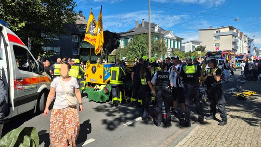 Auf einer Straße stehen mehere bunte Festwagen, eine Gruppe Polizisten, Feuerwehrfahrzeuge und einige Passanten