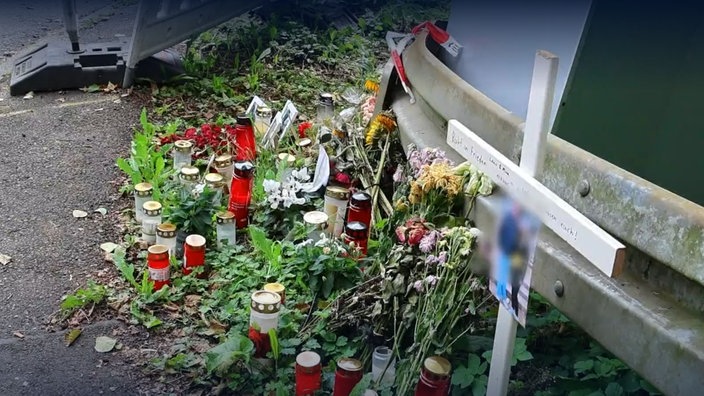 Blumen und Kerzen sind vor der Unfallstelle neben einer Leitplanke aufgestellt, auch ein Holzkreuz mit dem Foto der gestorbenen Geschwister steht an der Unfallstelle