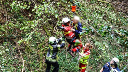 Eine alte Frau wird im Arm einer Feuerwehrmannes an einem Rettungsseil aus einem Waldstück herausgezogen.