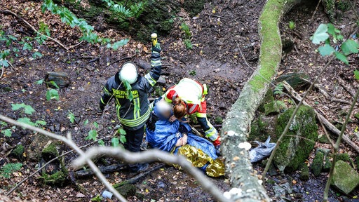 Eine Feuerwehrfrau legt eine Wärmedecke um eine Seniorin in einem Wald. Daneben winkt ihr männlicher Kollege in den Himmel.