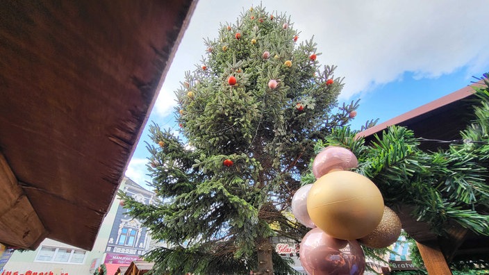Auf dem Foto ist ein mit Kugeln geschmückter Weihnachtsbaum zwischen Dächern von Weihnachtsmarkt-typischen Holzbuden.