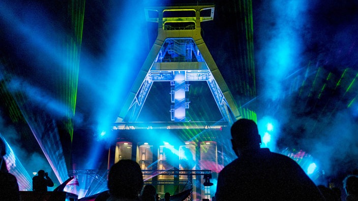 Tanzende Menschen inmitten einer Lasershow, im Hintergrund das Bergbau-Museum Bochum