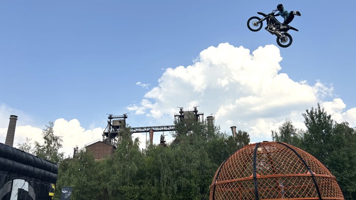 Ein Motorrad-Artist steht hoch in der Luft. Im Hintergrund sieht man Teile des Landschaftspark Duisburg-Nord.