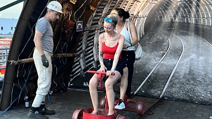Zwei Besucherinnen sitzen auf einem Grubenfahrrad. Ein Mann mit Schutzhelm schaut zu. 