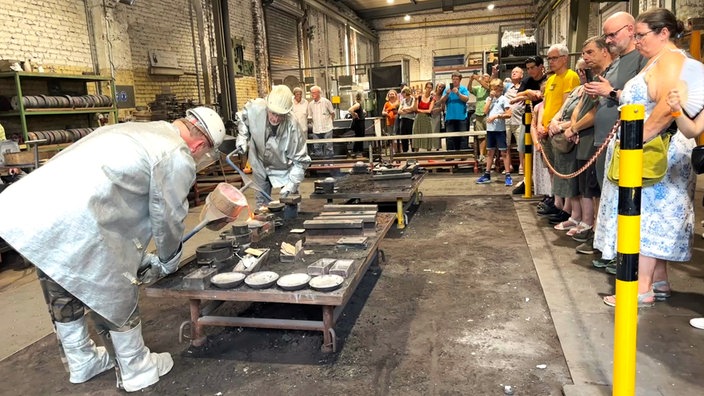 Zwei Männer gießen im LWL-Museum Henrichshütte in Hattingen flüssiges Aluminium in eine Form, Zuschauer stehen drumherum.