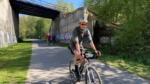 Ein Fahrradfahrer fährt lächelnd auf einer gut asphaltierten Bahntrasse im Grünen im Ruhrgebiet unter einer Brücke her