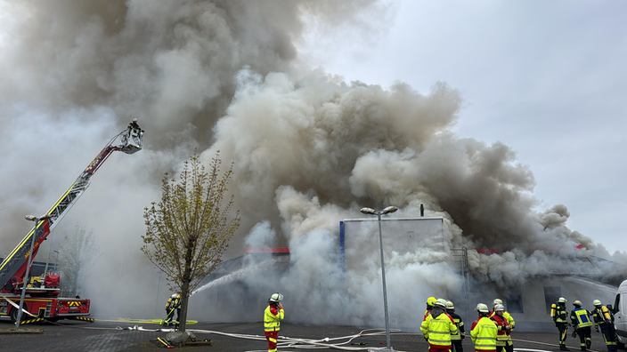 Die Feuerwehr ist mit einer Drehleiter vor Ort. Aus dem Gebäude steigen dichte, graue Rauchwolken empor. 