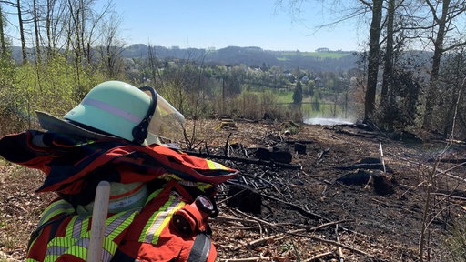 Das Bild zeigt einen Feuerwehrhelm vor der verbrannten Waldfläche.