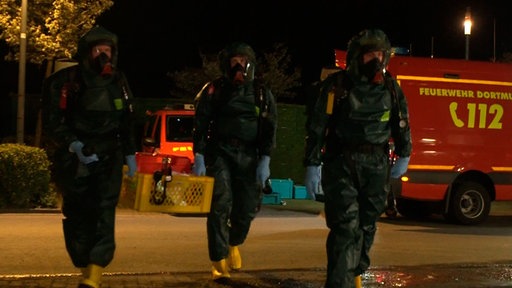Drei Personen in Schutzanzügen. Zwei davon tragen einen Korb in dem, Einsatzmaterialien liegen. 