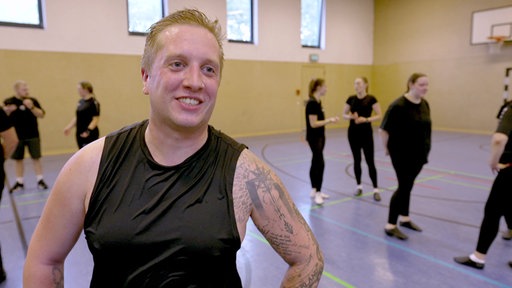 Blonder Mann in schwarzer Trainingskleidung steht lächelnd in einer Sporthalle