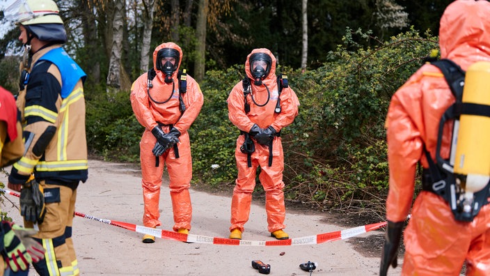 Einsatzkräfte in Schutzanzügen stehen in einem abgesperrten Bereich. 