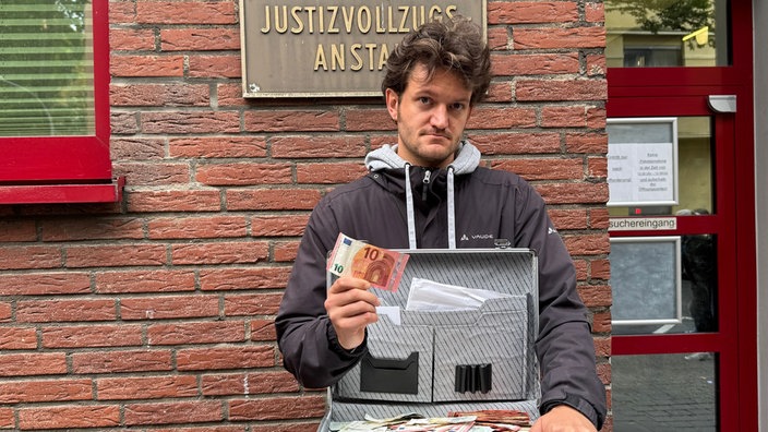 Mann blickt mit ernster Miene in die Kamera. Er hält einen offenen Koffer mit Geld in der Hand und einen zehn Euro Schein in die Höhe.