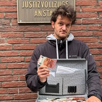 Mann blickt mit ernster Miene in die Kamera. Er hält einen offenen Koffer mit Geld in der Hand und einen zehn Euro Schein in die Höhe.