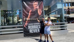 Zwei Männer stehen vor einem Robbie-Williams-Plakat.