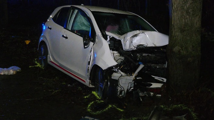 Ein weißes Auto ist nachts frontal gegen einen Baum geprallt und vorne stark beschädigt.