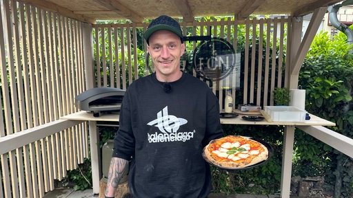Daniel Cyris lacht und holt eine frische Pizza aus dem kleinen Pizzaofen im Garten. 