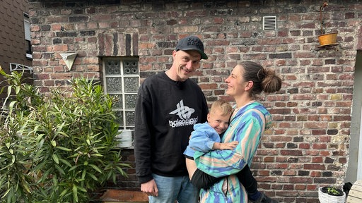 Daniel Cyris steht mit seiner Frau und seinem Sohn im Garten vor einer Backsteinmauer. Alle drei lachen.