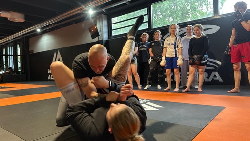 Daniel Cyris kämpft mit einer anderen Trainerin am Boden. Fünf Schülerinnen schauen zu.