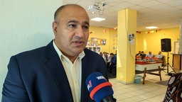 Ein Mann in dunkelblauem Anzug steht in einem großen Raum. Im Hintergrund sitzen mehrere Männer auf Stühlen
