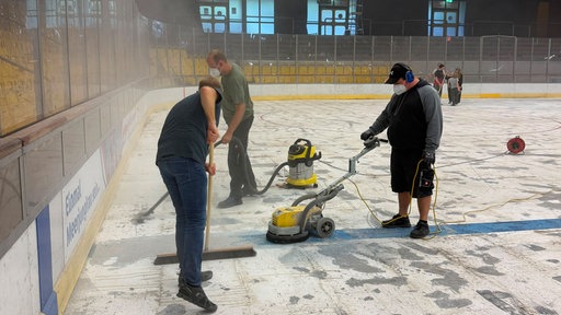 Drei Personen entfernen mit Maschinen und Werkzeugen die Eisfläche in einer Eishalle.