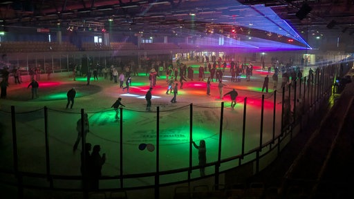 Mehrere Menschen laufen auf Schlittschuhen übers Eis in der Eishalle, die mit roten, grünen und lila Discolichtern erleuchtet ist