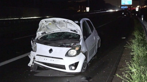 Ein weißer, verbeulter Twingo mit loser Motorhaube auf einer Autobahn bei Nacht
