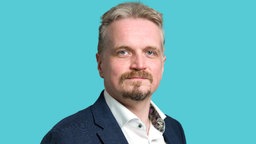 Ein Mann in weißem Hemd und dunkelblauem Jacket schaut vor hellblauem Hintergrund 