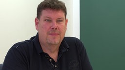 Ein Mann mit dunkelblauem Polo-Shirt sitzt auf einem Stuhl und schaut in die Kamera