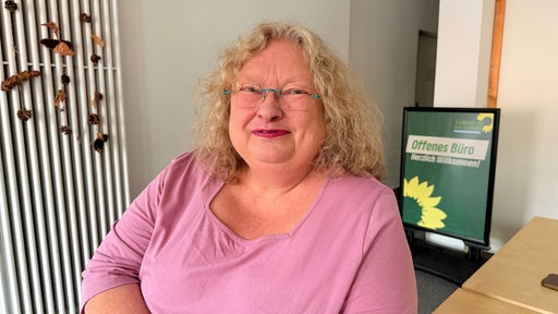 Auf dem Foto ist Anne Becker-Wischnewski, eine blonde Frau in einem pinken Langarmshirt. Sie sitzt in einem Grünen-Büro.