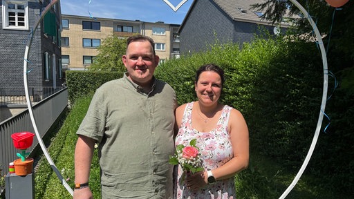 Ein Mann und eine Frau stehen im Garten umrahmt von einem silberfarbenem Metallherz und halten sich fest. Die Frau hält eine Rose in der Hand.