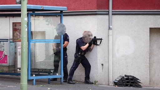 Polizisten an einer Hauswand. Einer hält eine Maschinenpistole im Anschlag.