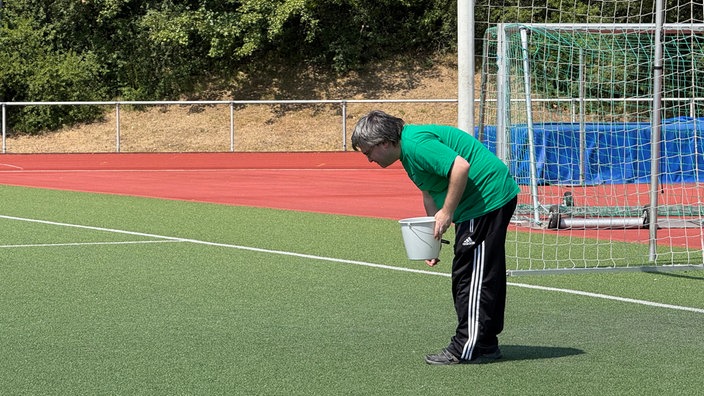 Ein Mann steht auf dem Rasen des Fußballplatzes, ist über den Boden gebäugt und sammelt Müll ein