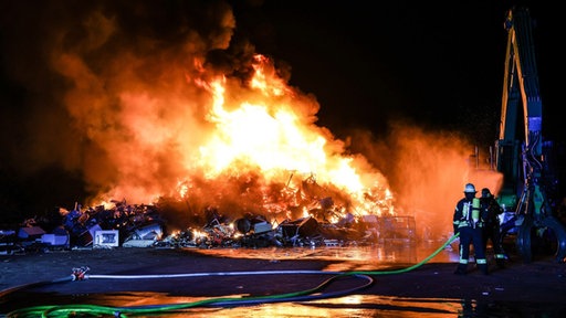 Ein großer Haufen Elektroschrott brennt, davor stehen Feuerwehrleute in Montur neben einer Drehleiter