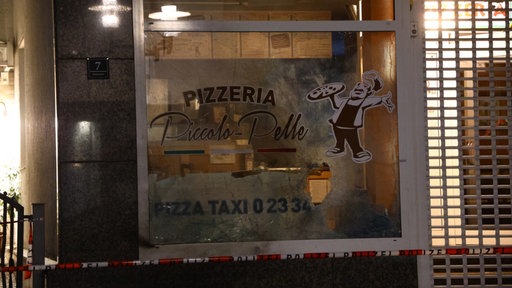 Die Außenansicht einer Pizzeria zeigt zerstörte Fensterscheiben, während der Bereich mit Polizeiband abgesperrt ist.
