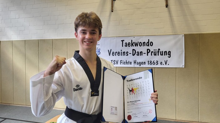 Maxim schaut in die Kamera und hält seine Urkunde für den schwarzen Gürtel im Teakwondo in der Hand.