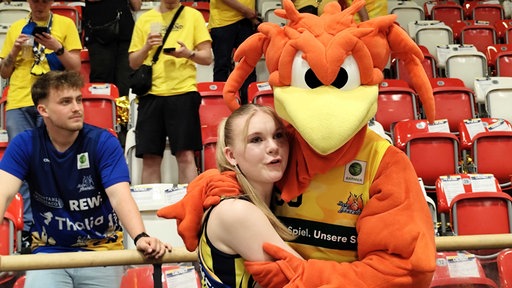 Eine junge blonde Frau, die in den Armen eines orangenen Plüschmaskottchens liegt, das böse guckt.