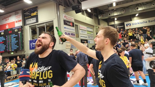 Ein blonder Mann, der von hinten einem dunkelhaarigen Bier überkippt. Beide tragen T-Shirts mit der Aufschrift "Aufsteiger".