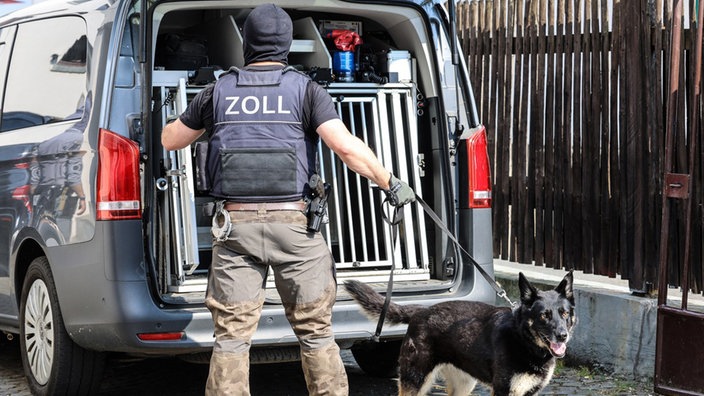 Ein Zoll-Beamter in Schutzweste öffnet eine Hundetransportbox in einem Auto. Er hält einen Hund an der Leine