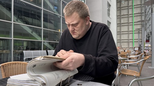 Ex-Häftling Manuel B. blättert durch dicken Ordner. Er sitzt in einem Außengastronomiebereich.