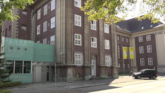 Ein aus roten Backsteinen gebautes Gebäude mit großen Fenstern umringt von grünen Bäumen.