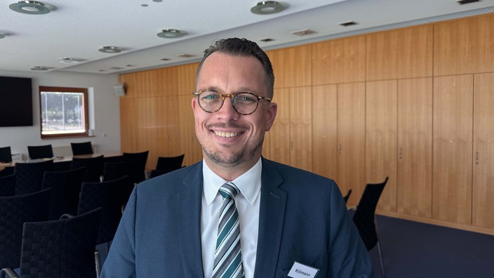 Ein Mann mit Brille, Anzug und zurückgegeelten Haaren. Er trägt ein Namensschild mit der Aufschrift "Kilimann".