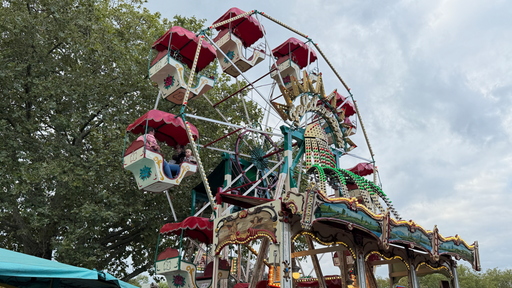 Ein Riesenrad aus dem Jahr 1902 steht auf einer Kirmes. Rote Überdachungen der Wagons sind zu erkennen.