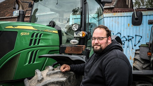 Landwirt Julian Freisendorf steht im schwarzen Pullover neben seinem grünen Traktor.