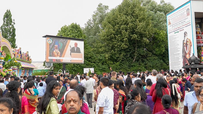 Eine große Gruppe Menschen steht unter freiem Himmel vor einer Leinwand, auf der Bundespräsident Frank-Walter Steinmeier hinter einem Rednerpult zu sehen ist.